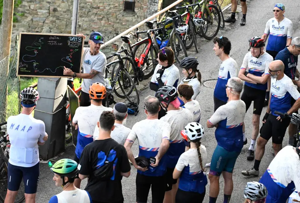 Un gruppo di ciclisti ascolta attentamente durante un briefing pre-uscita, con una lavagna che illustra il percorso e biciclette parcheggiate sullo sfondo.