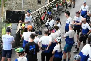 Un gruppo di ciclisti ascolta attentamente durante un briefing pre-uscita, con una lavagna che illustra il percorso e biciclette parcheggiate sullo sfondo.