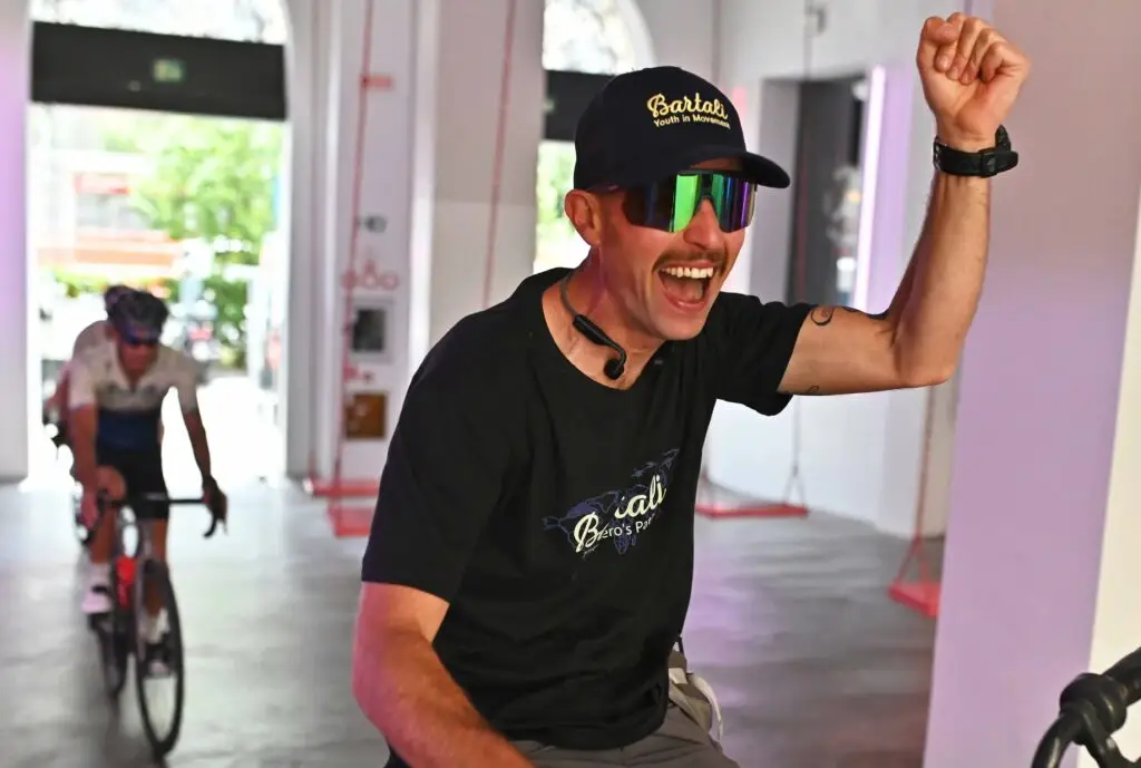 Ciclista sorridente con cappellino Bartali e occhiali da sole esulta, mentre un altro ciclista entra nella sala sullo sfondo.