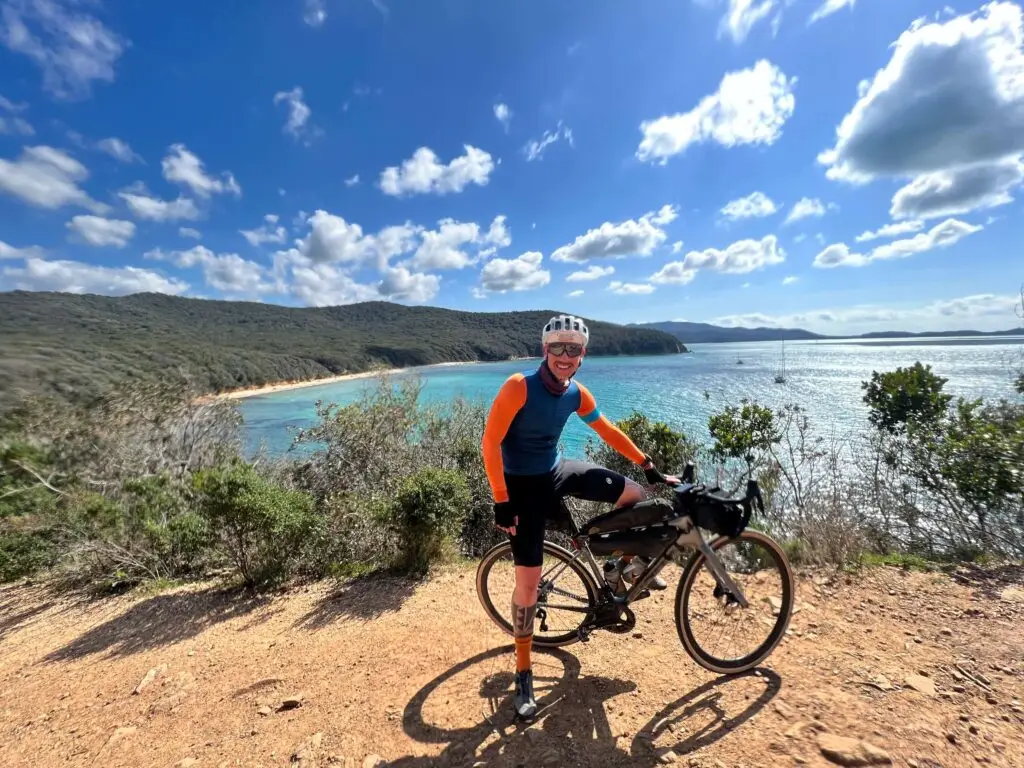 Un ciclista in abbigliamento tecnico posa con la sua bici da gravel su un sentiero sterrato, con alle spalle un panorama costiero di acque turchesi e colline verdi sull’isola d’Elba.