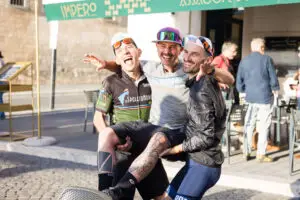 Tre ciclisti sorridenti in abbigliamento da gravel si abbracciano davanti a un bar, celebrando un momento di gioia e cameratismo dopo un'avventura in bicicletta
