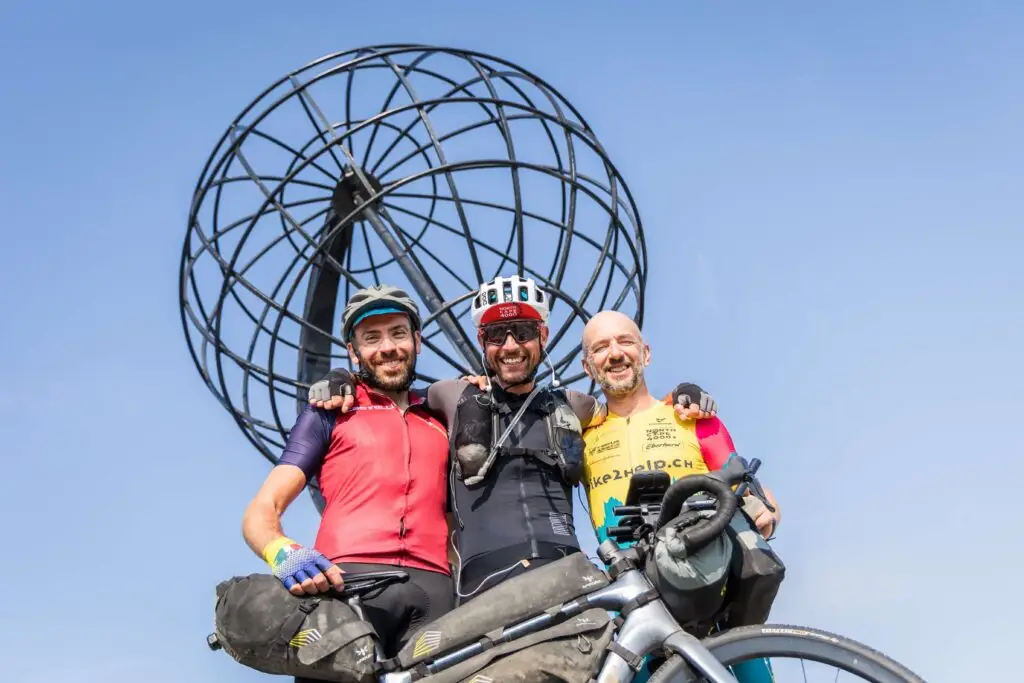 Tre ciclisti sorridenti con bici da bikepacking posano davanti al globo iconico di Capo Nord, sotto un cielo terso