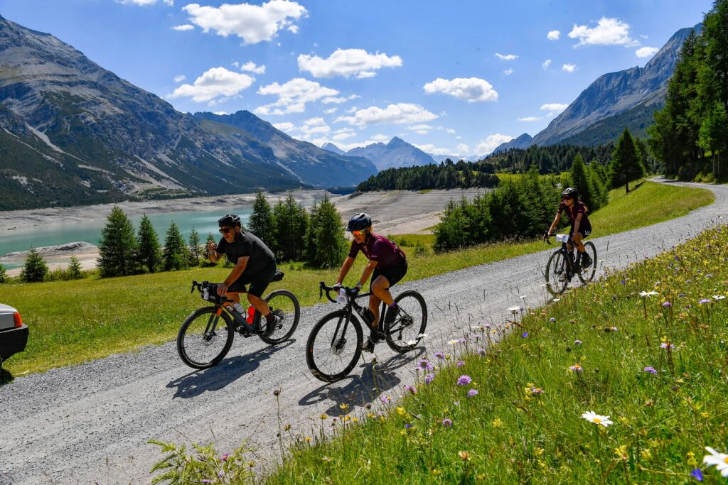 Tre ciclisti percorrono una strada sterrata immersa nella natura alpina, costeggiando un lago cristallino circondato da maestose montagne. I fiori selvatici e il cielo limpido completano questo scenario mozzafiato, simbolo di avventura e libertà.
