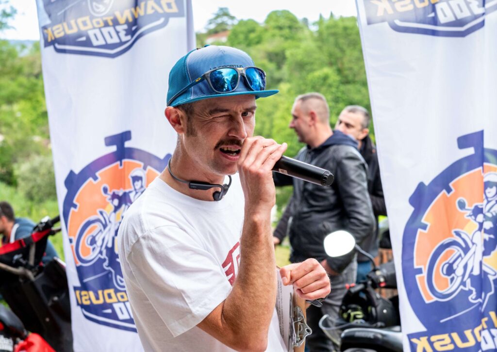 Un uomo con cappellino blu e occhiali da sole riflettenti parla al microfono durante l'evento motociclistico "Dawn to Dusk 300". Indossa una maglietta bianca e tiene in mano una cartelletta. Sullo sfondo ci sono due bandiere con il logo dell'evento raffigurante un motociclista stilizzato e delle motociclette parcheggiate.