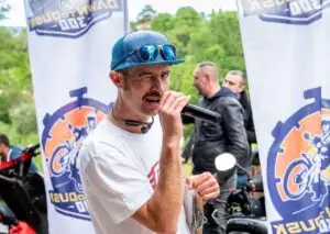 Un uomo con cappellino blu e occhiali da sole riflettenti parla al microfono durante l'evento motociclistico "Dawn to Dusk 300". Indossa una maglietta bianca e tiene in mano una cartelletta. Sullo sfondo ci sono due bandiere con il logo dell'evento raffigurante un motociclista stilizzato e delle motociclette parcheggiate.