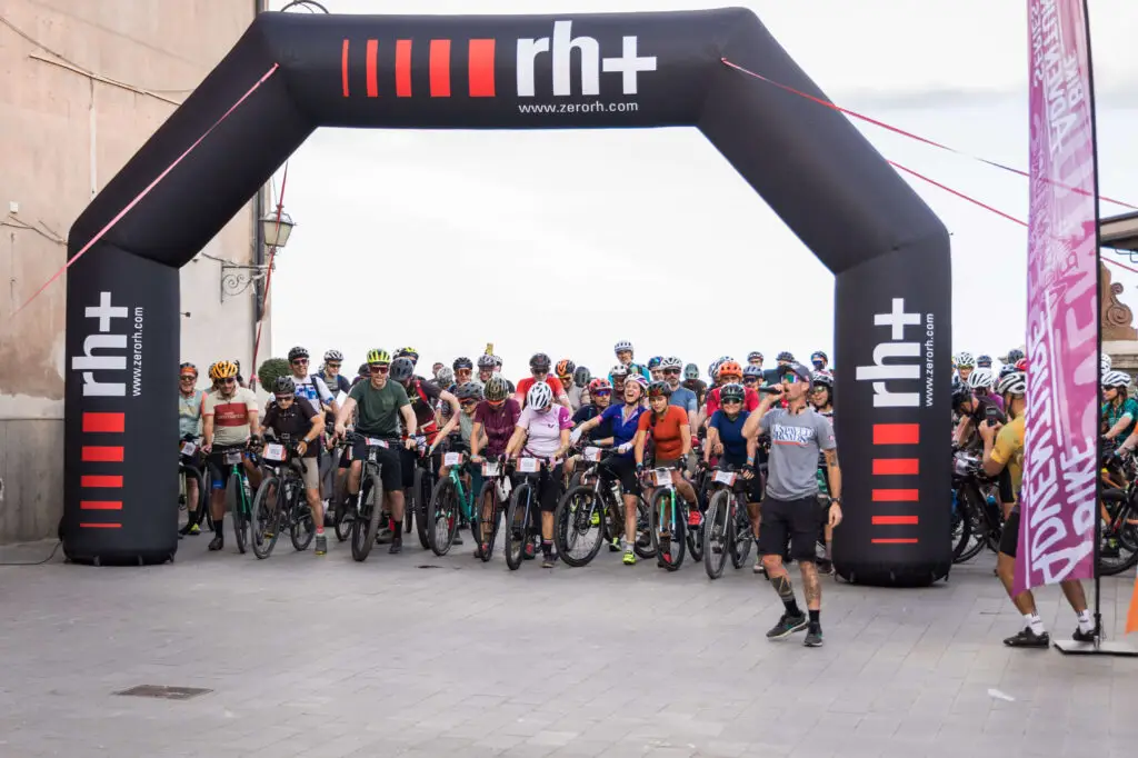 Ciclisti alla partenza di Unpaved Roads, radunati sotto un arco gonfiabile con il logo RH+ e una bandiera promozionale laterale. L'entusiasmo e la preparazione traspaiono nei volti dei partecipanti pronti a vivere un'avventura gravel unica.