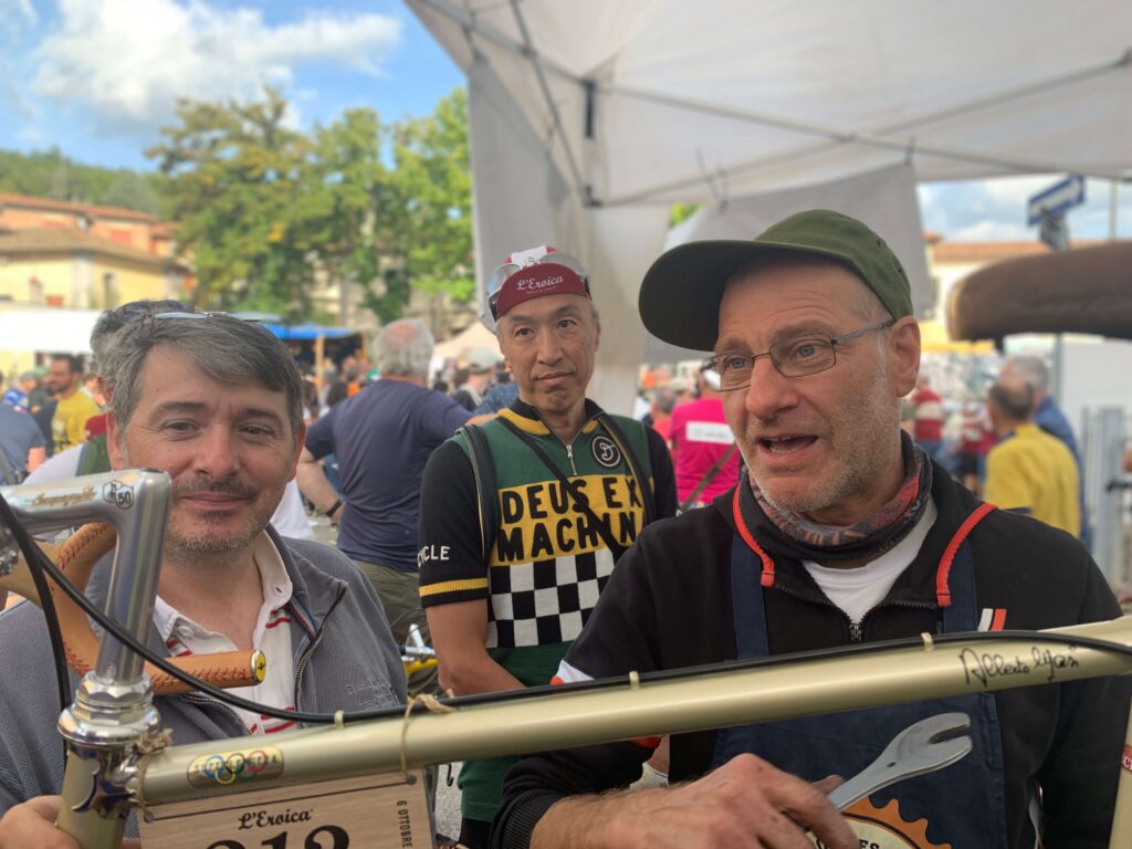 Meccanico esperto in una tenda all’Eroica mentre effettua la manutenzione di una bicicletta d’epoca, osservato da ciclisti appassionati