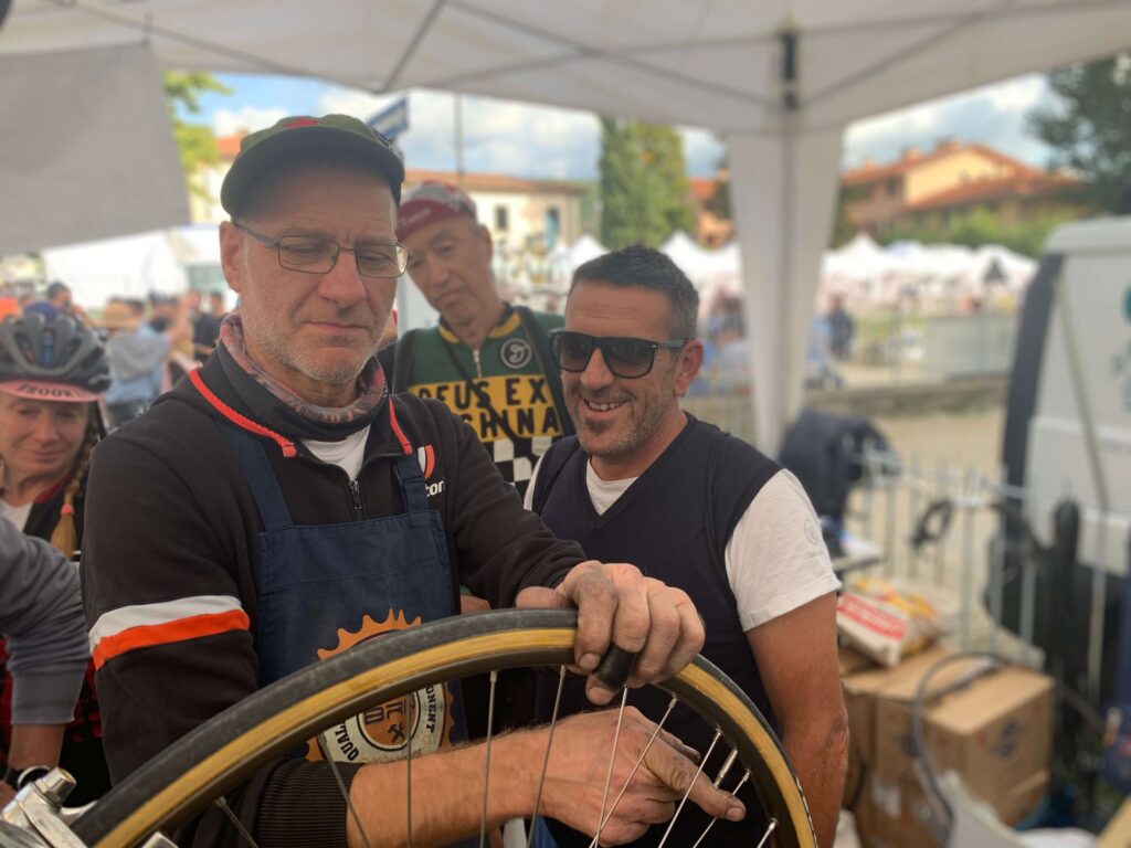 Meccanico con grembiule da officina al lavoro su una ruota, circondato da ciclisti durante un evento di biciclette d’epoca.