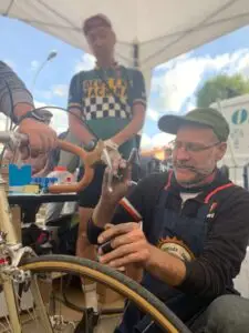 Meccanico esperto in una tenda all’Eroica mentre effettua la manutenzione di una bicicletta d’epoca, osservato da ciclisti appassionati.