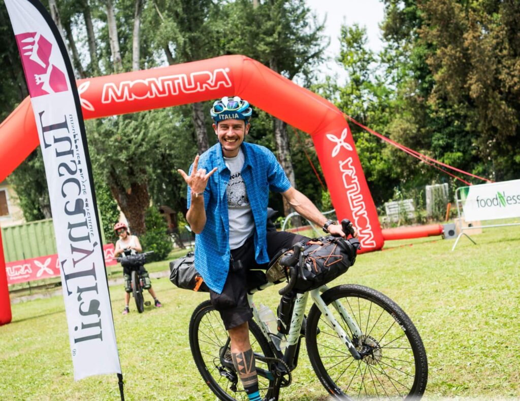 Un ciclista in assetto da bikepacking, con una camicia blu e casco tricolore, sorride e fa il segno “rock on” mentre taglia il traguardo dell’evento, sotto un arco gonfiabile Montura e accanto alla bandiera del Tuscany Trail.