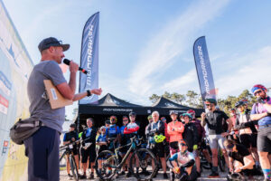 Un organizzatore parla al microfono durante il briefing di un evento gravel sponsorizzato Mondraker, davanti a un gruppo di ciclisti con bici e caschi, con bandiere e tende del brand sullo sfondo.