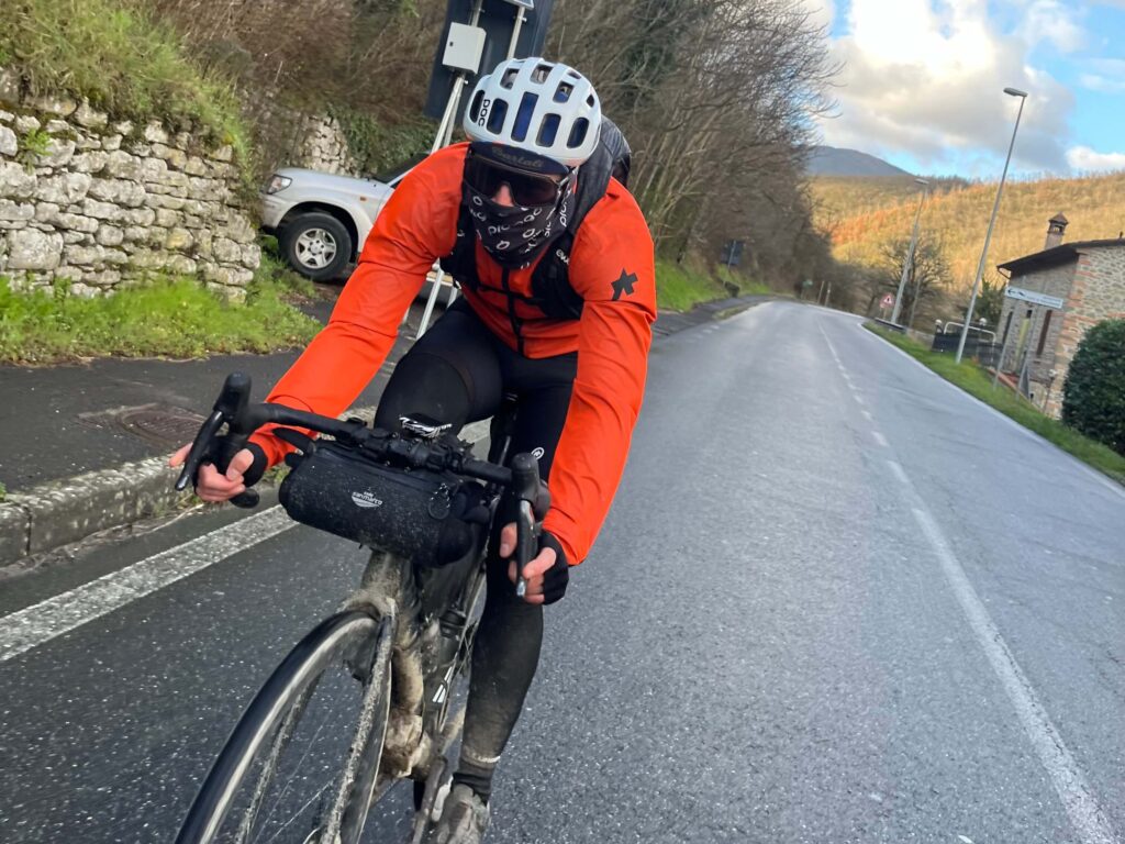 Ciclista con abbigliamento invernale che pedala su una strada bagnata in campagna, con borsa da manubrio e colline sullo sfondo.